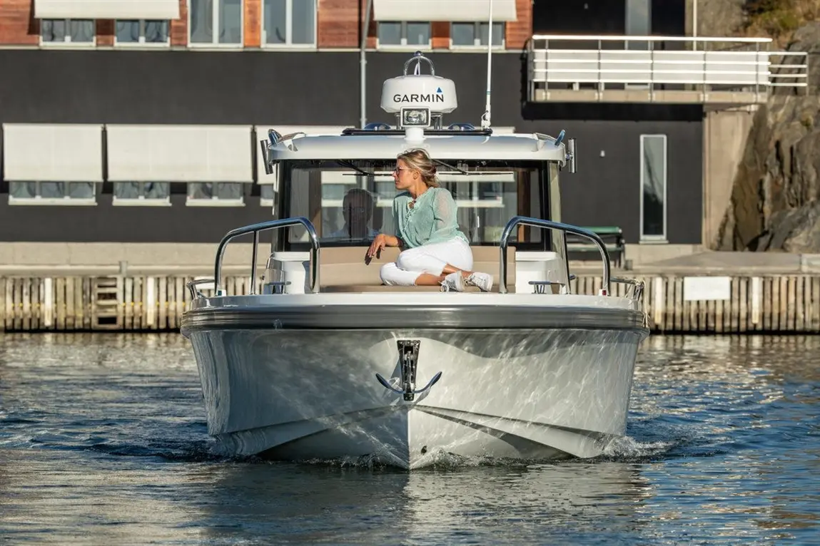 A woman in a light green top sits on a boat navigating calm waters near a modern dock and building, featuring Garmin equipment.